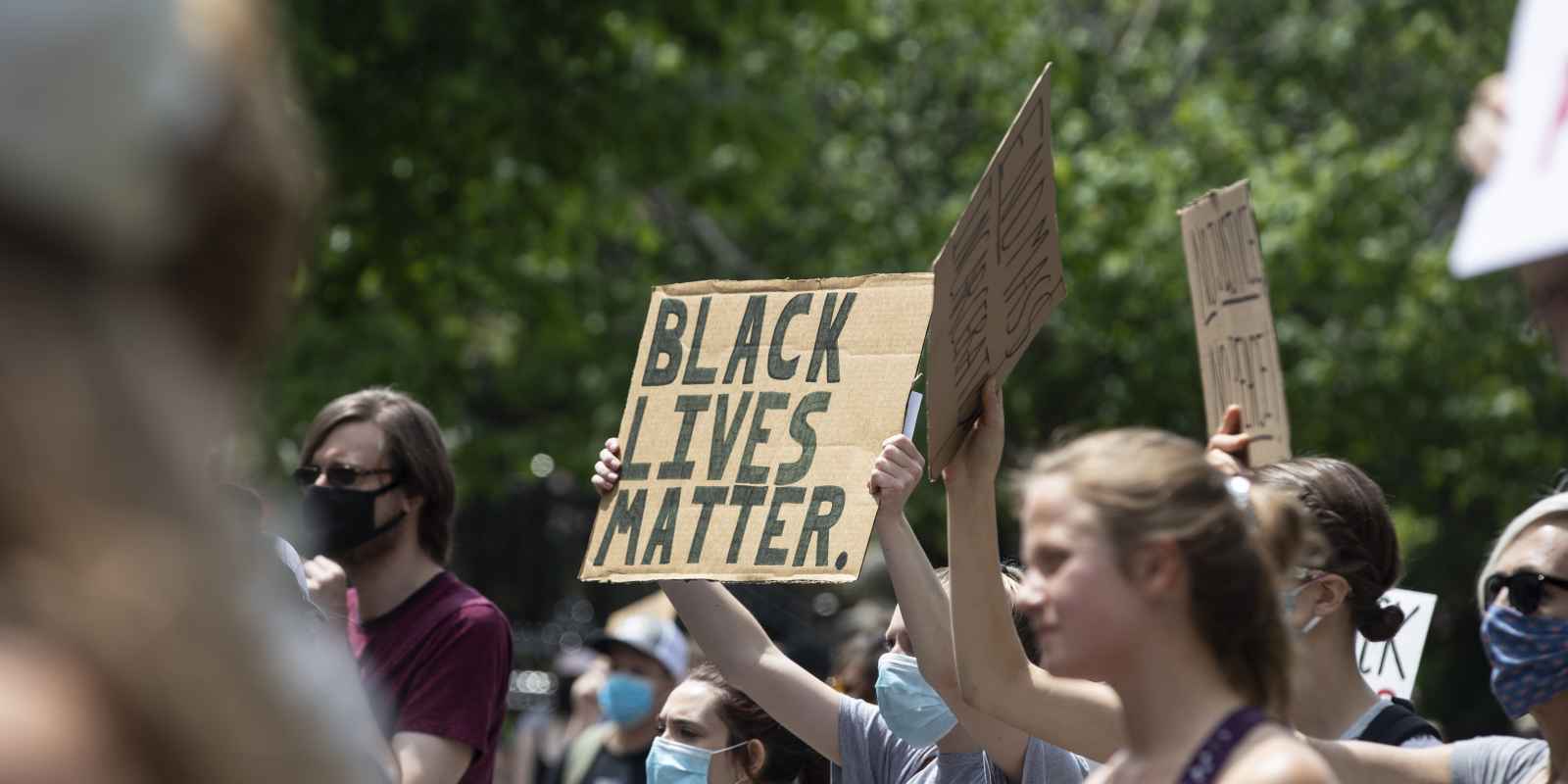 Photo of people protesting and holding homemade signs. The sign in the middle says, "Black Lives Matter."