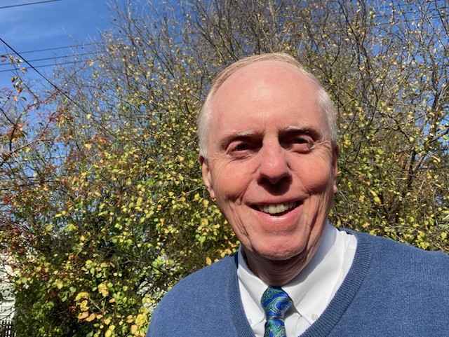 Photo of Bruce wearing a blue sweater and tie in front of a small, green tree.