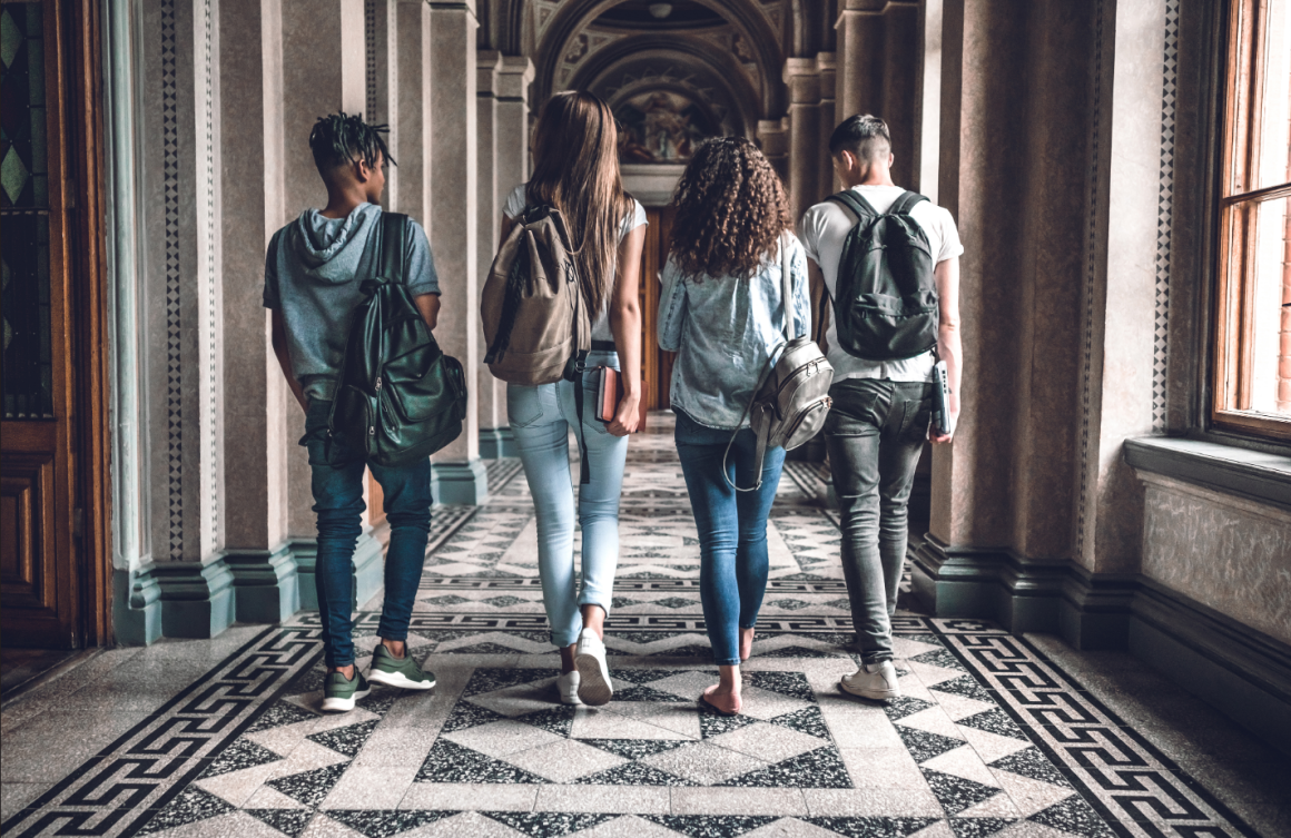 Photo of four college students wearing backpacks walking down a hallway with marble floor. 
