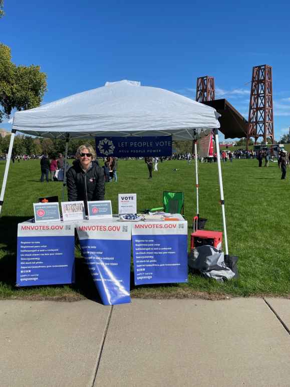 Photo of an individual with short blond hair, wearing sunglasses, standing under a white tent. In front of the person is a table with signs attached to it. The signs say, "MNvotes.gov." 