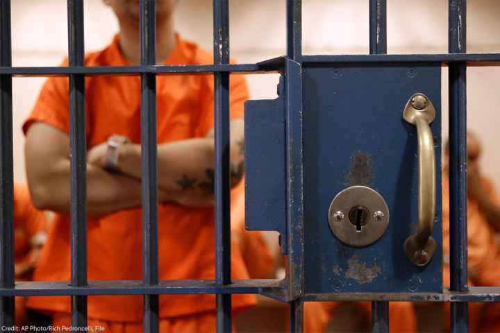 The lock and bars of a prison cell are at the forefront, while a detainee, standing in an orange jumpsuit and with his tattooed arms crossed waits in a cell with other detainees.