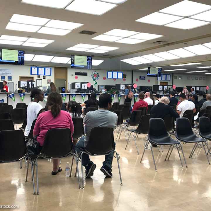 Long rows of chairs with people sitting inside the busy waiting room area of a DMV office.