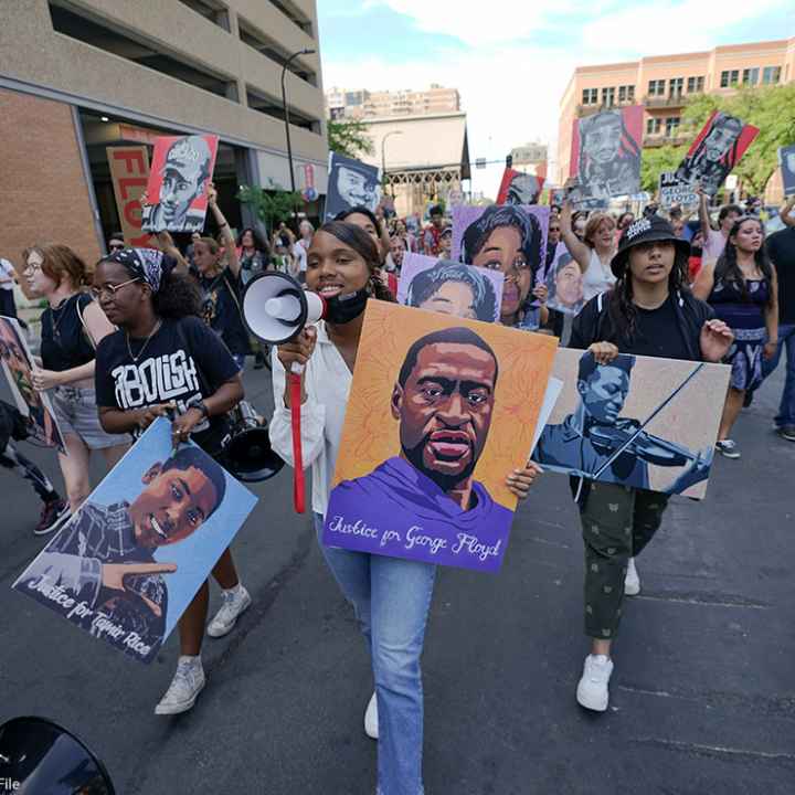 Protestors walking down the street, with one holding a microphone and a sign with art of George Floyd's face.